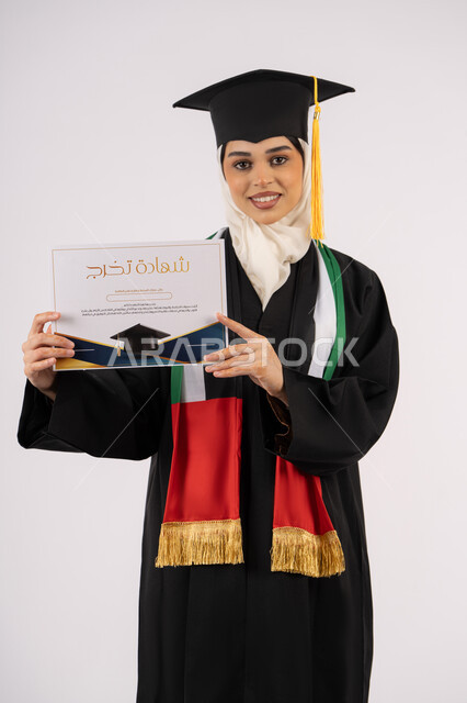 جودة التعليم في الامارات، الاحتفال بإنهاء الدراسة الجامعية، النظر الى الكاميرا بايماءات الفخر والسعادة بالوصول الى الحلم، بورتريه لخريجة عربية خليجية اماراتية محجبة ترتدي زي التخرج والقبعة تحمل بيدها شهادة التخرج، خلفية بيضاء