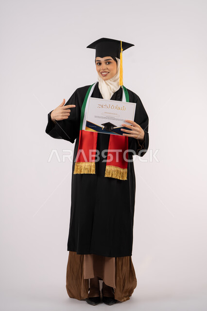 الإشارة بإصبع السبابة إلى شهادة التخرج، الاحتفال بإنهاء الدراسة الجامعية، بورتريه لخريجة عربية خليجية اماراتية محجبة ترتدي زي التخرج والقبعة تنظر الى الكاميرا بإيماءات الفخر والسعادة بالوصول الى الحلم، الجسم بالطول الكامل، خلفية بيضاء