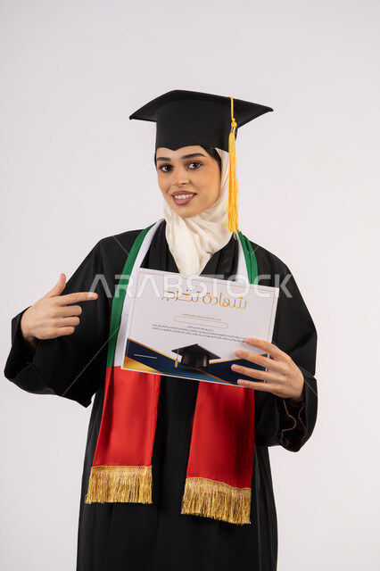 الاحتفال بإنهاء الدراسة الجامعية، الإشارة بإصبع السبابة إلى شهادة التخرج، بورتريه لخريجة عربية خليجية اماراتية محجبة ترتدي زي التخرج والقبعة تنظر الى الكاميرا بإيماءات الفخر والسعادة بالوصول الى الحلم، خلفية بيضاء