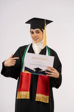 الاحتفال بإنهاء الدراسة الجامعية، الإشارة بإصبع السبابة إلى شهادة التخرج، بورتريه لخريجة عربية خليجية اماراتية محجبة ترتدي زي التخرج والقبعة تنظر الى الكاميرا بإيماءات الفخر والسعادة بالوصول الى الحلم، خلفية بيضاء