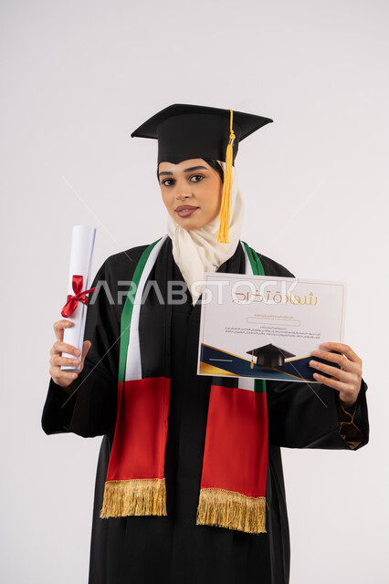 إيماءات الفخر والسعادة بالوصول الى الحلم، تحقيق الأحلام والطموحات، الاحتفال بإنهاء الدراسة الجامعية، بورتريه لخريجة عربية خليجية اماراتية محجبة ترتدي زي التخرج والقبعة تحمل وثيقة التخرج وتنظر الى الكاميرا، خلفية بيضاء