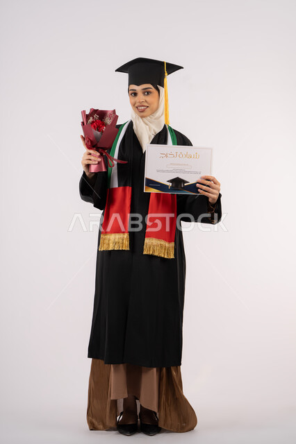 النظر إلى الكاميرا بتعابير السرور، الاحتفال بإنهاء الدراسة الجامعية، بورتريه لخريجة عربية خليجية اماراتية محجبة ترتدي زي التخرج والقبعة تمسك بيدها بوكيه ورد والشهادة بإيماءات السعادة بالوصول لحلم النجاح،، الجسم بالطول الكامل، خلفية بيضاء
