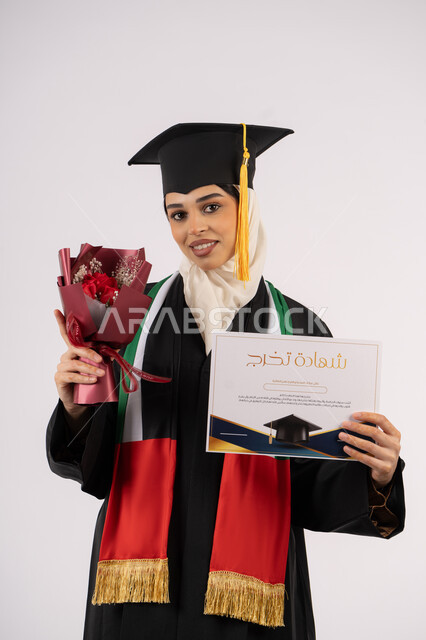 أهمية الدراسة الجامعية، النظر إلى الكاميرا بتعابير السرور، مناسبة واحتفال جامعي، بورتريه لخريجة عربية خليجية اماراتية محجبة ترتدي زي التخرج والقبعة تمسك بيدها بوكيه ورد والشهادة بإيماءات السعادة بالوصول لحلم النجاح، خلفية بيضاء