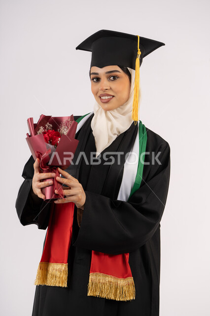 مشاعر الفرح والسعادة بالوصول لحلم النجاح، بورتريه لخريجة عربية خليجية اماراتية محجبة ترتدي زي التخرج والقبعة تمسك بيدها بوكيه ورد تنظر إلى الكاميرا بتعابير السرور، الاحتفال بإنهاء الدراسة الجامعية، خلفية بيضاء