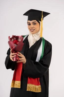 مشاعر الفرح والسعادة بالوصول لحلم النجاح، بورتريه لخريجة عربية خليجية اماراتية محجبة ترتدي زي التخرج والقبعة تمسك بيدها بوكيه ورد تنظر إلى الكاميرا بتعابير السرور، الاحتفال بإنهاء الدراسة الجامعية، خلفية بيضاء