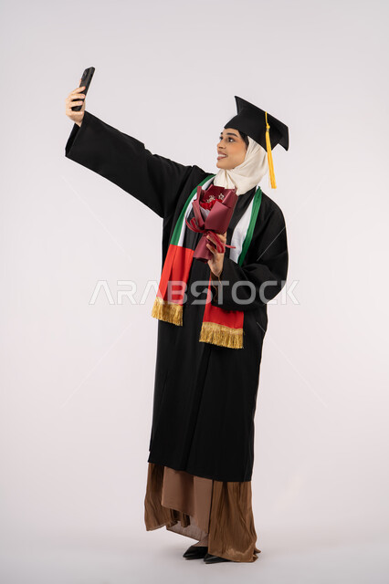 استخدام التقنية الحديثة، الاحتفال بإنهاء الدراسة الجامعية، تقديم الورد في المناسبات، الاحتفاظ بالذكريات السعيدة، بورتريه لخريجة عربية خليجية اماراتية ترتدي عباءه وقبعة التخرج تقوم بالتقاط صورة سيلفي تذكارية، الجسم بالطول الكامل، خلفية بيضاء