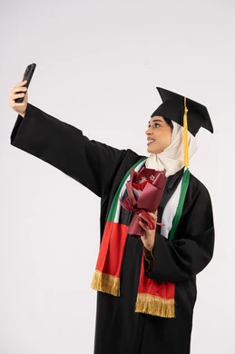 الاحتفاظ بالذكريات السعيدة، استخدام التقنية الحديثة، الاحتفال بإنهاء الدراسة الجامعية، تقديم الورد في المناسبات، بورتريه لخريجة عربية خليجية اماراتية ترتدي عباءه وقبعة التخرج تقوم بالتقاط صورة سيلفي تذكارية، خلفية بيضاء