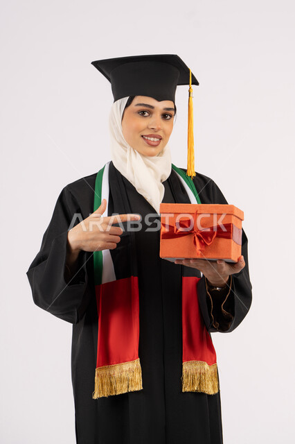 Giving gifts on occasions, celebrating the completion of university studies, gestures of happiness for reaching the dream of success, portrait of a veiled Arab Gulf Emirati graduate wearing a graduation gown and cap, pointing with her index finger to the gift box, white background