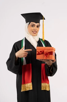 تقديم الهدايا في المناسبات، الاحتفال بإنهاء الدراسة الجامعية، إيماءات السعادة بالوصول لحلم النجاح، بورتريه لخريجة عربية خليجية اماراتية محجبة ترتدي زي التخرج والقبعة تشير بإصبع السبابة إلى صندوق الهدية، خلفية بيضاء