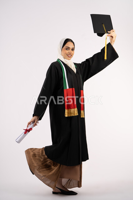 المناسبات والاحتفالات الجامعية، النظر الى الكاميرا بايماءات الفخر، الوصول إلى حلم النجاح، الاحتفال بإنهاء الدراسة الجامعية، بورتريه لخريجة عربية خليجية اماراتية محجبة ترتدي زي التخرج تمسك بيدها القبعة ووثيقة التفوق، صورة الجسم بالكامل، خلفية بيضاء