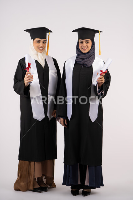 مشاعر الفرح بتحقيق الأحلام، الاحتفال مع الأصدقاء بالتخرج من الجامعة، النظر إلى الكاميرا بإيماءات السعادة والفخر، بورتريه لخريجتين جامعيتين عربيتين خليجيتين اماراتيتين ترتديان ملابس التخرج وتمسكان وثيقة التفوق، صورة لكامل الجسم، خلفية بيضاء