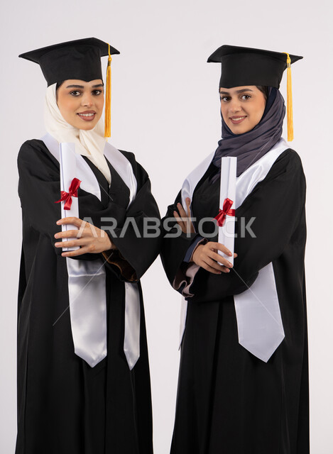 جودة التعليم في الامارات، الفرح بتحقيق الأحلام، الاحتفال مع الأصدقاء بالتخرج من الجامعة، النظر إلى الكاميرا بإيماءات السعادة والفخر، بورتريه لخريجتين جامعيتين عربيتين خليجيتين اماراتيتين ترتديان ملابس التخرج تقفان بأيدي متقاطعة وتمسكان وثيقة التفوق، خلفية بيضاء