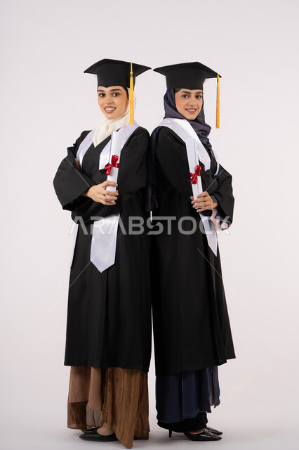 جودة التعليم في جامعات الامارات العربية، النظر إلى الكاميرا بإيماءات السعادة والفخر، الاحتفال مع الأصدقاء بالتخرج من الجامعة، بورتريه لخريجتين جامعيتين عربيتين خليجيتين اماراتيتين ترتديان ملابس التخرج وتمسكان شهادة النجاح والتفوق، طول الجسم بالكامل، خلفية بيضاء