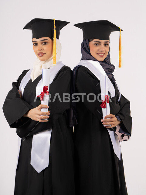 التعليم في جامعات الامارات العربية، الاحتفال بالتخرج من الجامعة، بورتريه لخريجتين جامعيتين عربيتين خليجيتين اماراتيتين ترتديان ملابس التخرج وتمسكان شهادة النجاح والتفوق، ايماءات السعادة والفرح والفخر، خلفية بيضاء