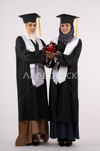 مناسبة واحتفال جامعي، مفهوم الاجتهاد والتفوق، النظر إلى الكاميرا بإيماءات السعادة والفخر، الاحتفال مع الأصدقاء بالتخرج من الجامعة، بورتريه لخريجتين جامعيتين عربيتين خليجيتين اماراتيتين ترتديان ملابس التخرج وتمسكان باقة ورد، طول الجسم بالكامل، خلفية بيضاء