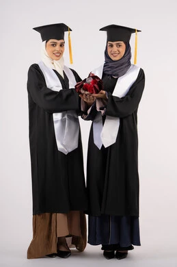 University occasion and celebration, concept of diligence and excellence, looking at the camera with gestures of happiness and pride, celebrating university graduation with friends, portrait of two Arab Gulf Emirati university graduates wearing graduation clothes and holding a bouquet of flowers, full body length, white background