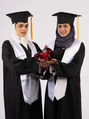 Celebrating university graduation with friends, university occasion and celebration, concept of hard work and excellence, looking at the camera with gestures of happiness and pride, portrait of two Arab Gulf Emirati university graduates wearing graduation clothes and holding a bouquet of flowers, white background
