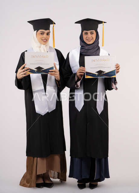 مفهوم الاجتهاد والتفوق، النظر إلى الكاميرا بإيماءات السعادة والفخر، الاحتفال مع الأصدقاء بالتخرج من الجامعة، بورتريه لخريجتين جامعيتين عربيتين خليجيتين اماراتيتين ترتديان ملابس التخرج وتمسكان شهادة النجاح، طول الجسم بالكامل، خلفية بيضاء