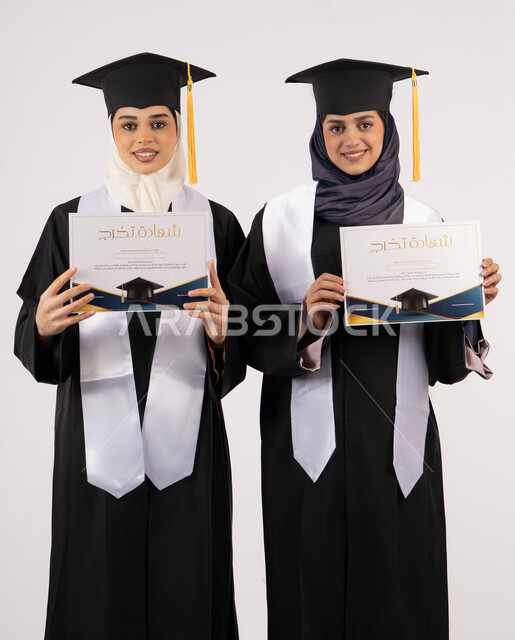 النظر إلى الكاميرا بإيماءات السعادة والفرح والفخر، التعليم في جامعات الامارات العربية، الاحتفال مع الأصدقاء بالتخرج من الجامعة، بورتريه لخريجتين جامعيتين عربيتين خليجيتين اماراتيتين ترتديان ملابس التخرج وتمسكان شهادة النجاح والتفوق، خلفية بيضاء
