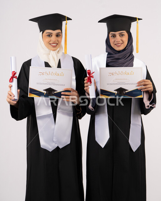 Education in UAE universities, university graduation celebration, portrait of two Emirati Arab Gulf university graduates wearing graduation attire and holding certificates of success and excellence, gestures of happiness, joy and pride, white background