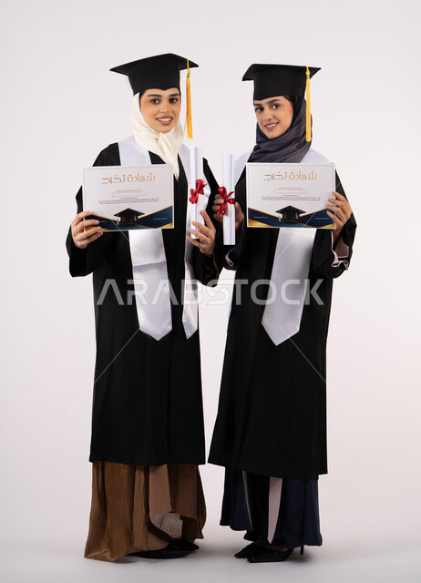Quality of education in UAE universities, looking at the camera with gestures of happiness and pride, celebrating university graduation with friends, portrait of two Emirati Arab Gulf university graduates wearing graduation clothes and holding their certificates of success and excellence, full body length, white background