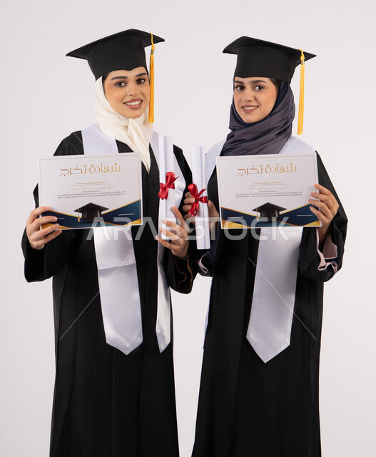 النظر إلى الكاميرا بإيماءات السعادة والفرح والفخر، التعليم في جامعات الامارات العربية، الاحتفال مع الأصدقاء بالتخرج من الجامعة، بورتريه لخريجتين جامعيتين عربيتين خليجيتين اماراتيتين ترتديان ملابس التخرج وتمسكان شهادة النجاح والتفوق، خلفية بيضاء