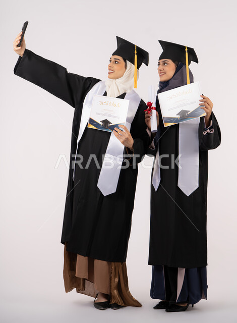 The joy of receiving a graduation certificate, using modern technology, celebrating the completion of university studies, preserving happy memories, a portrait of an Arab Gulf Emirati female graduate wearing an abaya and graduation cap, taking a commemorative selfie with her friend, full body, white background