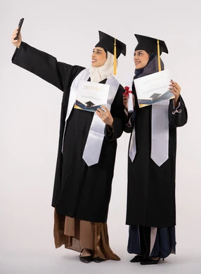 فرحة الحصول على شهادة التخرج، استخدام التقنية الحديثة، الاحتفال بإنهاء الدراسة الجامعية،الاحتفاظ بالذكريات السعيدة، بورتريه لخريجة عربية خليجية اماراتية ترتدي عباءه وقبعة التخرج تقوم بالتقاط صورة سيلفي تذكارية برفقة صديقتها، الجسم بالطول الكامل، خلفية بيضاء