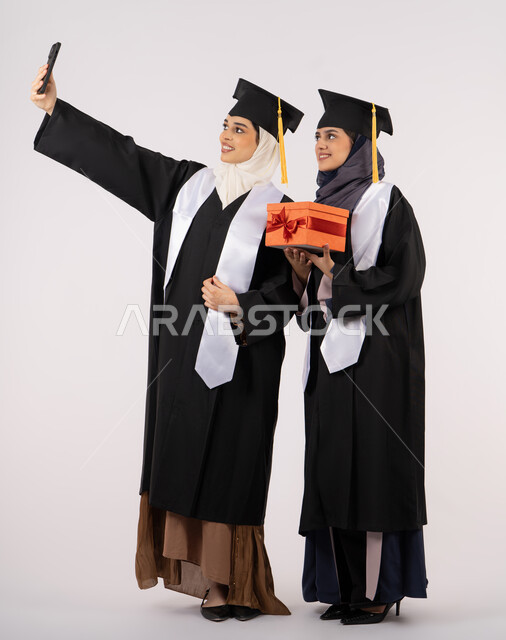 استخدام التقنية الحديثة، الاحتفال بإنهاء الدراسة الجامعية، تقديم الهدايا في المناسبات، الاحتفاظ بالذكريات السعيدة، بورتريه لخريجة عربية خليجية اماراتية ترتدي عباءه وقبعة التخرج تقوم بالتقاط صورة سيلفي تذكارية برفقة صديقتها، الجسم بالطول الكامل، خلفية بيضاء