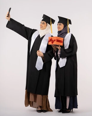 استخدام التقنية الحديثة، الاحتفال بإنهاء الدراسة الجامعية، تقديم الهدايا في المناسبات، الاحتفاظ بالذكريات السعيدة، بورتريه لخريجة عربية خليجية اماراتية ترتدي عباءه وقبعة التخرج تقوم بالتقاط صورة سيلفي تذكارية برفقة صديقتها، الجسم بالطول الكامل، خلفية بيضاء