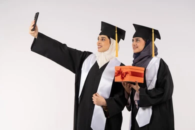 الاحتفاظ بالذكريات السعيدة، تقديم الهدايا في المناسبات، الاحتفال بإنهاء الدراسة الجامعية، استخدام التقنية الحديثة، مفهوم الاجتهاد والتفوق، بورتريه لخريجة عربية خليجية اماراتية ترتدي عباءه وقبعة التخرج تقوم بالتقاط صورة سيلفي تذكارية برفقة صديقتها، خلفية بيضاء