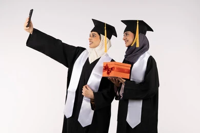 تقديم الهدايا في المناسبات، الاحتفاظ بالذكريات السعيدة، الاحتفال بإنهاء الدراسة الجامعية، استخدام التقنية الحديثة، مفهوم الاجتهاد والتفوق، بورتريه لخريجة عربية خليجية اماراتية ترتدي عباءه وقبعة التخرج تقوم بالتقاط صورة سيلفي تذكارية برفقة صديقتها، خلفية بيضاء