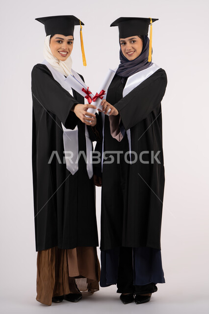 مفهوم الاجتهاد والتفوق، النظر إلى الكاميرا بإيماءات السعادة والفخر، الاحتفال مع الأصدقاء بالتخرج من الجامعة، بورتريه لخريجتين جامعيتين عربيتين خليجيتين اماراتيتين ترتديان ملابس التخرج وتمسكان شهادة النجاح، طول الجسم بالكامل، خلفية بيضاء