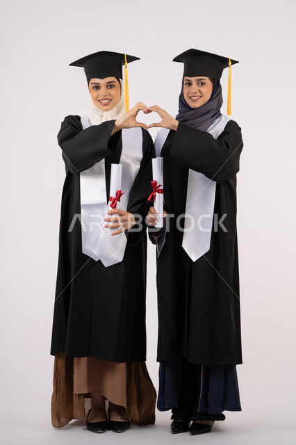 تشكيل اليدين على شكل قلب، الوقوف باستقامة والنظر إلى الكاميرا بإيماءات السعادة والفخر، الاحتفال مع الأصدقاء بالتخرج من الجامعة، بورتريه لخريجتين جامعيتين عربيتين خليجيتين اماراتيتين ترتديان ملابس التخرج وتمسكان شهادة النجاح، طول الجسم بالكامل، خلفية بيضاء