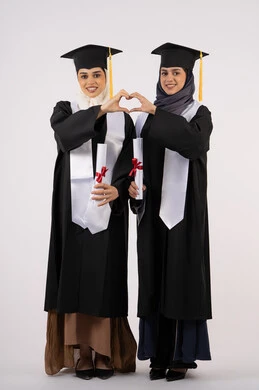 تشكيل اليدين على شكل قلب، الوقوف باستقامة والنظر إلى الكاميرا بإيماءات السعادة والفخر، الاحتفال مع الأصدقاء بالتخرج من الجامعة، بورتريه لخريجتين جامعيتين عربيتين خليجيتين اماراتيتين ترتديان ملابس التخرج وتمسكان شهادة النجاح، طول الجسم بالكامل، خلفية بيضاء