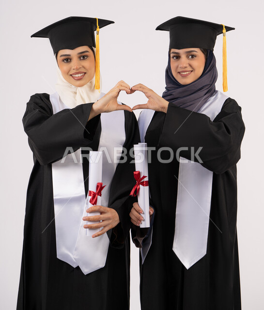 Feelings of joy at success, hands forming a heart shape, standing straight and looking at the camera with gestures of happiness and pride, celebrating university graduation with friends, portrait of two Arab Gulf Emirati university graduates wearing graduation clothes and holding their certificates of excellence, white background