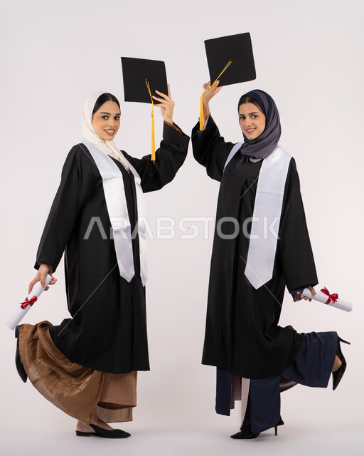 رفع قبعة التخرج للأعلى، مفهوم الاجتهاد والتفوق، النظر إلى الكاميرا بإيماءات السعادة والفخر، الاحتفال مع الأصدقاء بالتخرج من الجامعة، بورتريه لخريجتين جامعيتين عربيتين خليجيتين اماراتيتين ترتديان زي التخرج وتمسكان شهادة النجاح، طول الجسم بالكامل، خلفية بيضاء