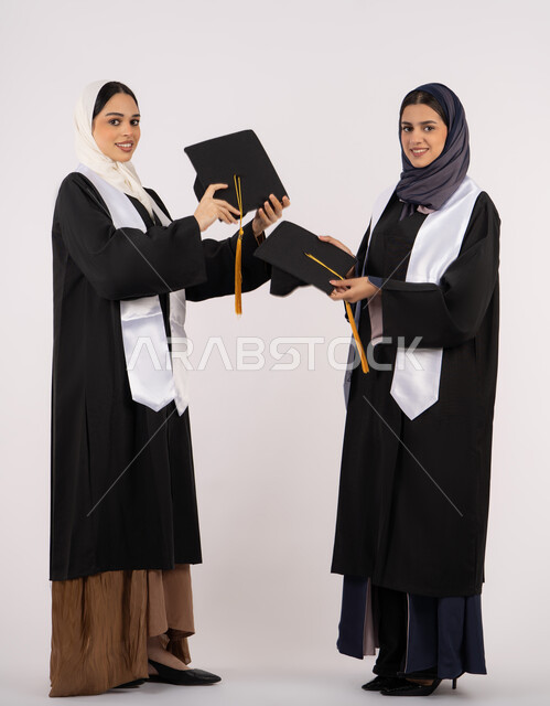 النجاح والاجتهاد والتفوق، النظر إلى الكاميرا بإيماءات السعادة والفخر، الاحتفال مع الأصدقاء بالتخرج من الجامعة، بورتريه لخريجتين جامعيتين عربيتين خليجيتين اماراتيتين ترتديان زي التخرج وتمسكان قبعة التخرج، طول الجسم بالكامل، خلفية بيضاء