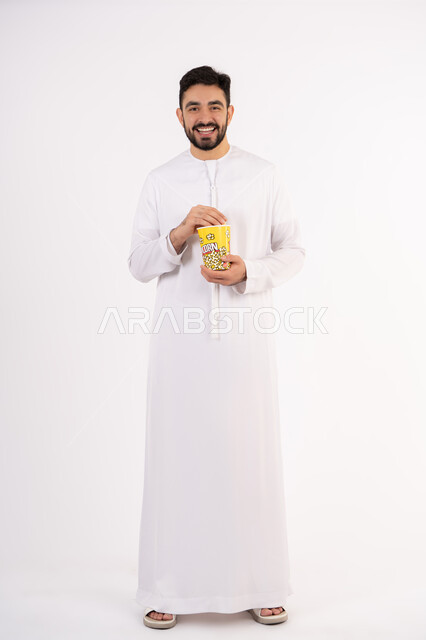 مفهوم التسليه والترفيه، تعابير المرح والاستمتاع ، الاستمتاع بتناول الفوشار، قضاء أوقات ممتعة ومسلية، بورتريه لرجل عربي خليجي اماراتي يرتدي الزى التقليدي يحمل بيده علبة الفشار ويقوم بمشاهده الافلام ، مشاهدة فيلم سينمائي ، خلفيه بيضاء