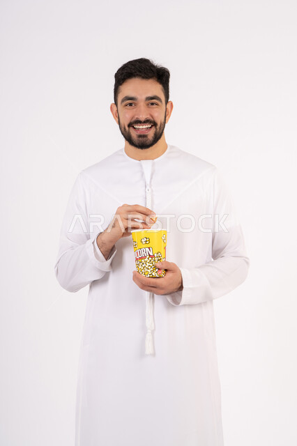الاستمتاع بتناول الفوشار، مفهوم التسليه والترفيه، تعابير المرح والاستمتاع ، قضاء أوقات ممتعة ومسلية، بورتريه لرجل عربي خليجي اماراتي يرتدي الزى التقليدي يحمل بيده علبة الفشار ويقوم بمشاهده الافلام ، مشاهدة فيلم سينمائي ، خلفيه بيضاء