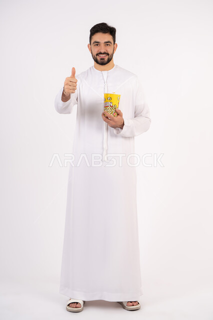 Expressions of quality, excellence and satisfaction, raising the index finger in a gesture of approval, having a good and entertaining time, the concept of entertainment and recreation, a portrait of an Emirati Gulf Arab man wearing traditional clothing, holding a box of popcorn in his hand and watching movies, watching a movie, white background