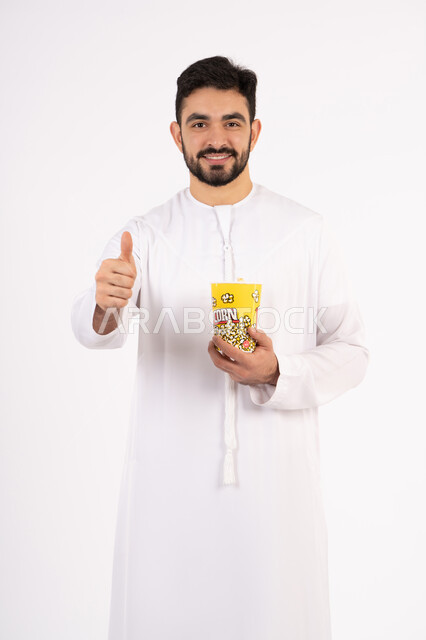 رفع اصبع السبابه بايماءات الموافقه ، تعابير الجودة والامتياز والرضي ، قضاء أوقات ممتعة ومسلية، مفهوم التسليه والترفيه، بورتريه لرجل عربي خليجي اماراتي يرتدي الزى التقليدي يحمل بيده علبة الفشار ويقوم بمشاهده الافلام ، مشاهدة فيلم سينمائي ، خلفيه بيضاء