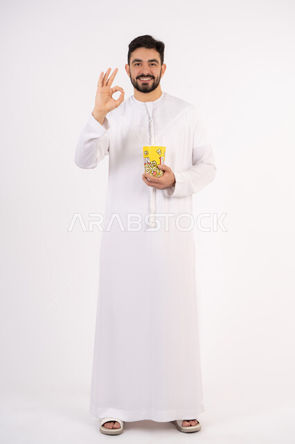 قضاء أوقات ممتعة ومسلية، تعابير الجودة والامتياز والرضي ،  رفع اليد بايماءات الموافقه ، مفهوم التسليه والترفيه، بورتريه لرجل عربي خليجي اماراتي يرتدي الزى التقليدي يحمل بيده علبة الفشار ويقوم بمشاهده الافلام ، مشاهدة فيلم سينمائي ، خلفيه بيضاء