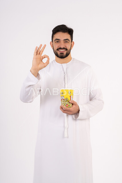 مشاهدة فيلم سينمائي ،قضاء أوقات ممتعة ومسلية، تعابير الجودة والامتياز والرضي ، رفع اليد بايماءات الموافقه ، مفهوم التسليه والترفيه، بورتريه لرجل عربي خليجي اماراتي يرتدي الزى التقليدي يحمل بيده علبة الفشار ويقوم بمشاهده الافلام ، خلفيه بيضاء