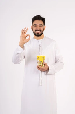 مشاهدة فيلم سينمائي ،قضاء أوقات ممتعة ومسلية، تعابير الجودة والامتياز والرضي ، رفع اليد بايماءات الموافقه ، مفهوم التسليه والترفيه، بورتريه لرجل عربي خليجي اماراتي يرتدي الزى التقليدي يحمل بيده علبة الفشار ويقوم بمشاهده الافلام ، خلفيه بيضاء