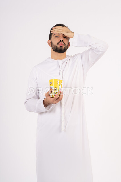 The concept of regretting something, watching a movie, having a good time, the concept of entertainment and amusement, an Arab Gulf Emirati man wearing traditional clothing holding a box of popcorn and watching movies, white background