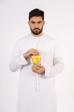 Watching a movie, angry facial and hand gestures, having a good time, the concept of entertainment, an Emirati Gulf Arab man wearing traditional clothing holding a popcorn box and watching movies, white background