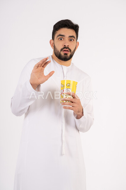Raising hand with shock gestures, expressions of fear and horror, concept of entertainment and amusement, portrait of an Emirati Gulf Arab man wearing a kandura and ghutra watching movies, holding a cup full of popcorn, watching movies and theater shows in the cinema, having a good time eating popcorn while watching TV, white background