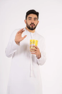 رفع اليد بايماءات الصدمه، تعابير الخوف والرعب ، مفهوم التسليه والترفيه ، بورتريه لرجل عربي خليجي امارتي يرتدي الكندوره والغتره يشاهد الافلام ، يحمل بيده كوب مليئ بالبوب كورن، مشاهدة الأفلام والعروض المسرحية في السينما، قضاء وقت ممتع بتناول الفشار اثناء مشاهده التلفاز ، خلفيه بيضاء