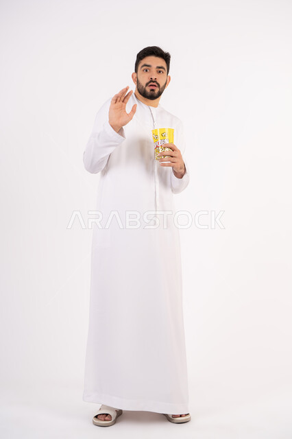 تعابير الخوف والرعب ، مفهوم التسليه والترفيه ، بورتريه لرجل عربي خليجي امارتي يرتدي الكندوره والغتره يشاهد الافلام ، يحمل بيده كوب مليئ بالبوب كورن، مشاهدة الأفلام والعروض المسرحية في السينما، قضاء وقت ممتع بتناول الفشار اثناء مشاهده التلفاز ، خلفيه بيضاء